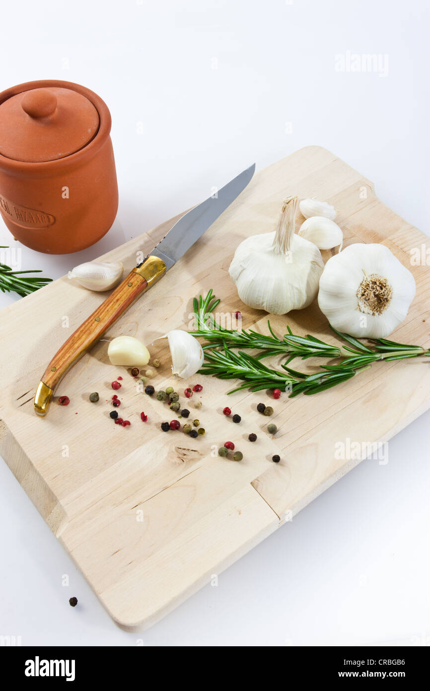 Knoblauch (Allium Sativum) auf einem Holzbrett mit Messer, Römertopf, Rosmarin und schwarzem und rotem Pfeffer Stockfoto