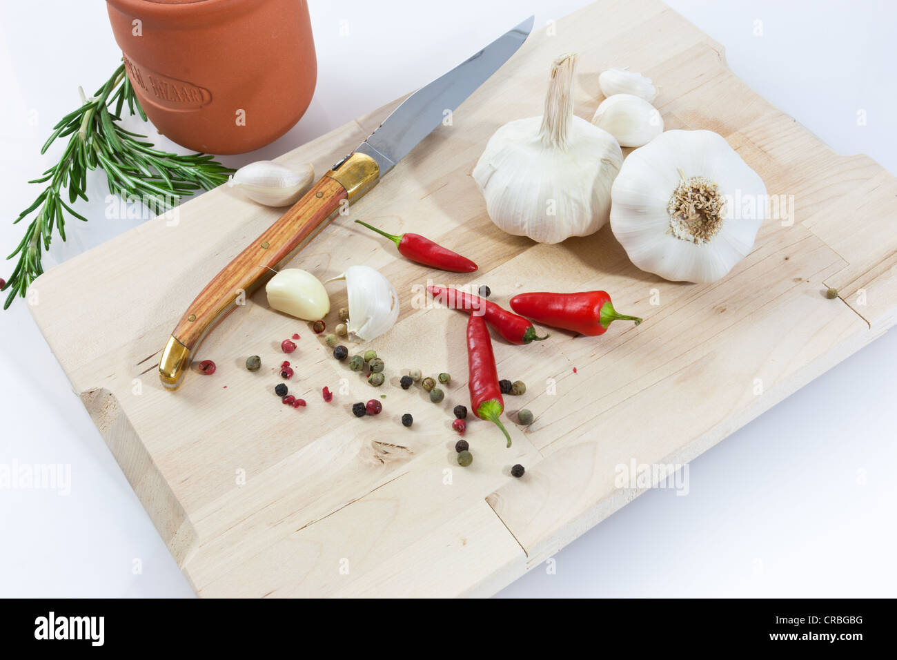Knoblauch (Allium Sativum) auf einem Holzbrett mit Messer, Tontopf, Chili und schwarzem und rotem Pfeffer Stockfoto