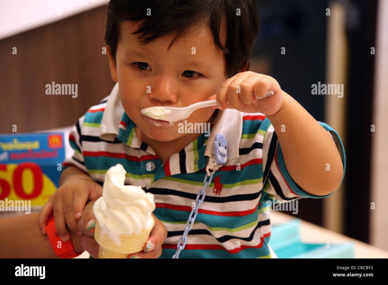 Es ist ein Foto eines niedlichen chinesischen Jungen eine Vanille-Eis mit einem Löffel zu essen. Er trägt einen Polo mit mehrfarbigen Streifen. Er liebt es Stockfoto