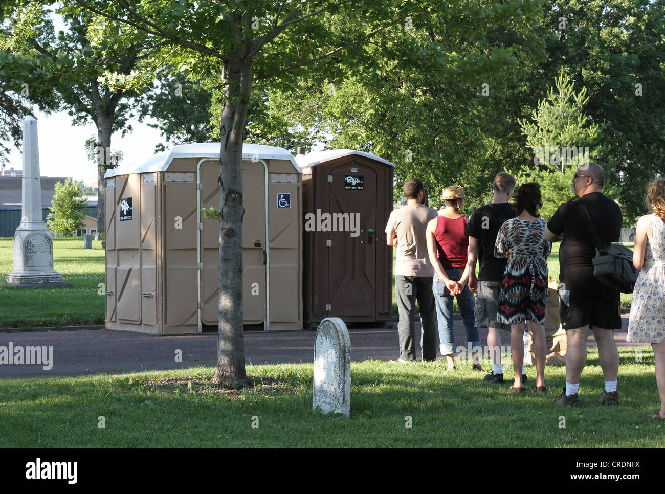 Leute standen Schlange, um die tragbare Badezimmer auf einem Friedhof zu verwenden. Stockfoto