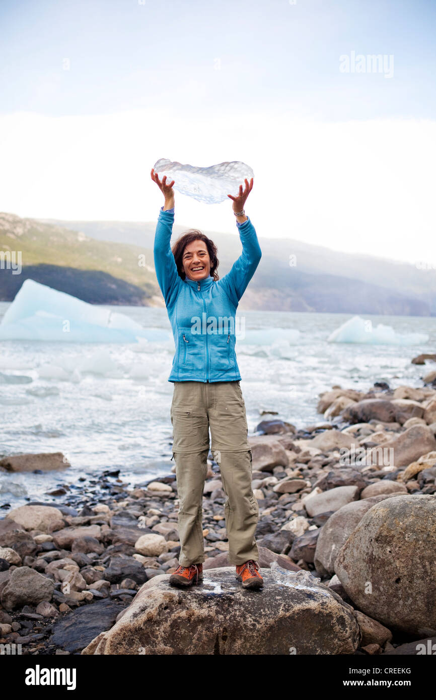 Eine lächelnde Frau hält ein Stück Eis, das von einem Gletscher auf das felsige Ufer stehend fiel. Stockfoto