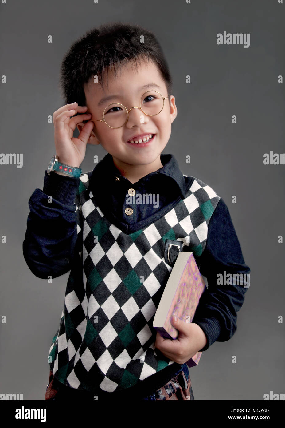 5 Jahre alten chinesischen Jungen tragen von trendigen Getriebe, stehend, seine Brille anpassen und ein Buch hält Stockfoto