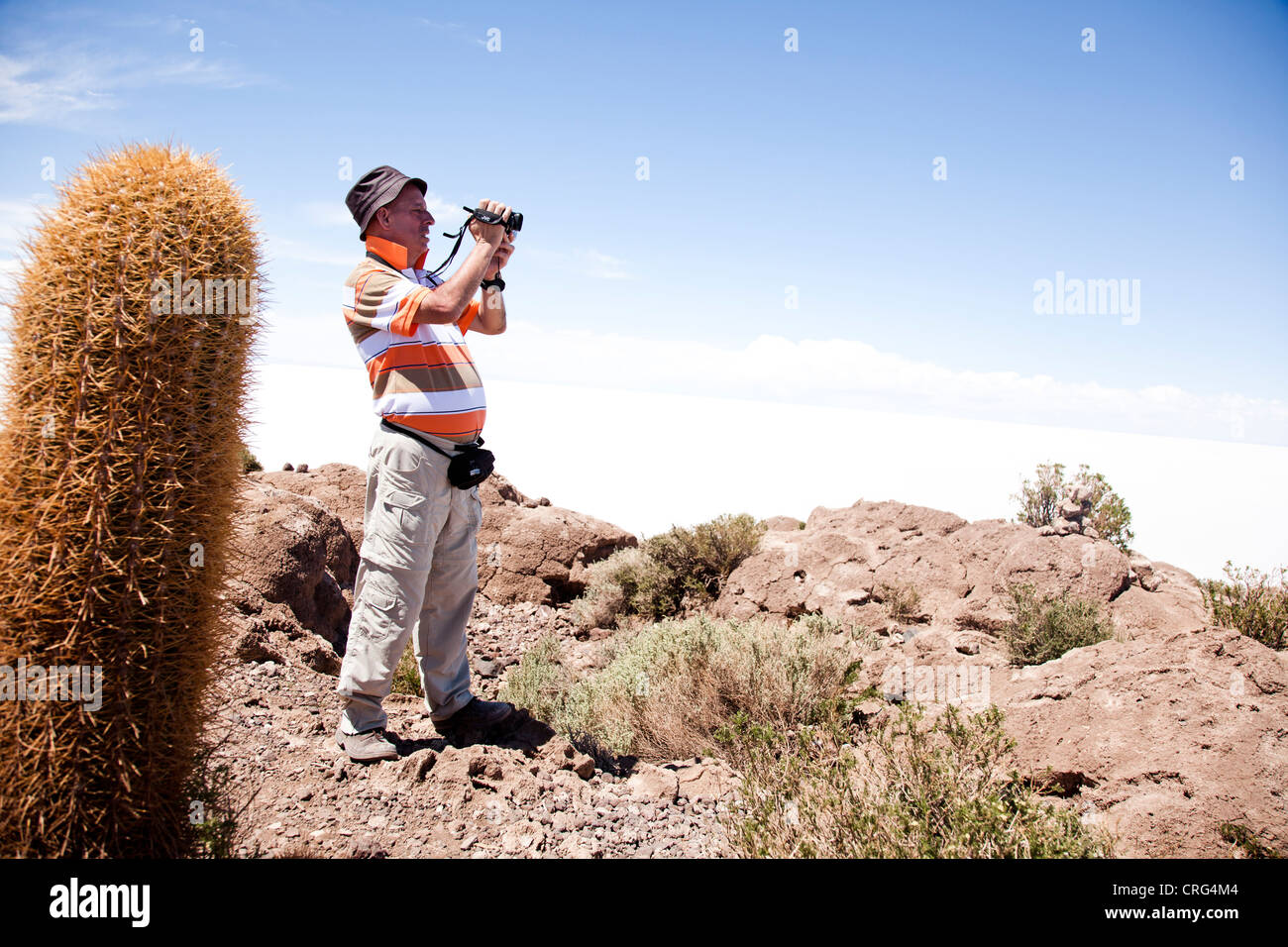 Ein Mann in einem Schlapphut mit touristischen Kleidung an schießt eine Video auf einer felsigen Landschaft mit einem Kaktus im Vordergrund. Stockfoto