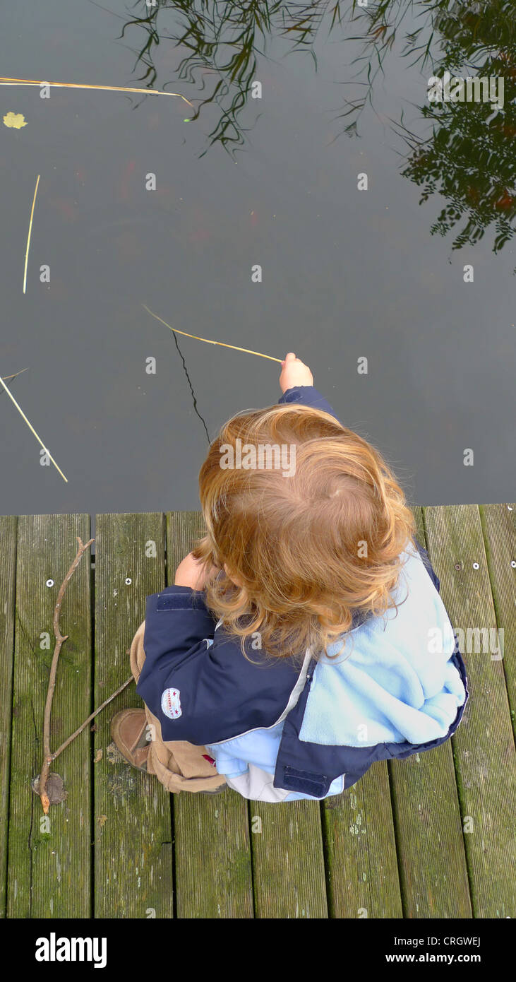 Kind sitzt auf einem Holzsteg, der einen Speer ins Wasser eintauchen Stockfoto