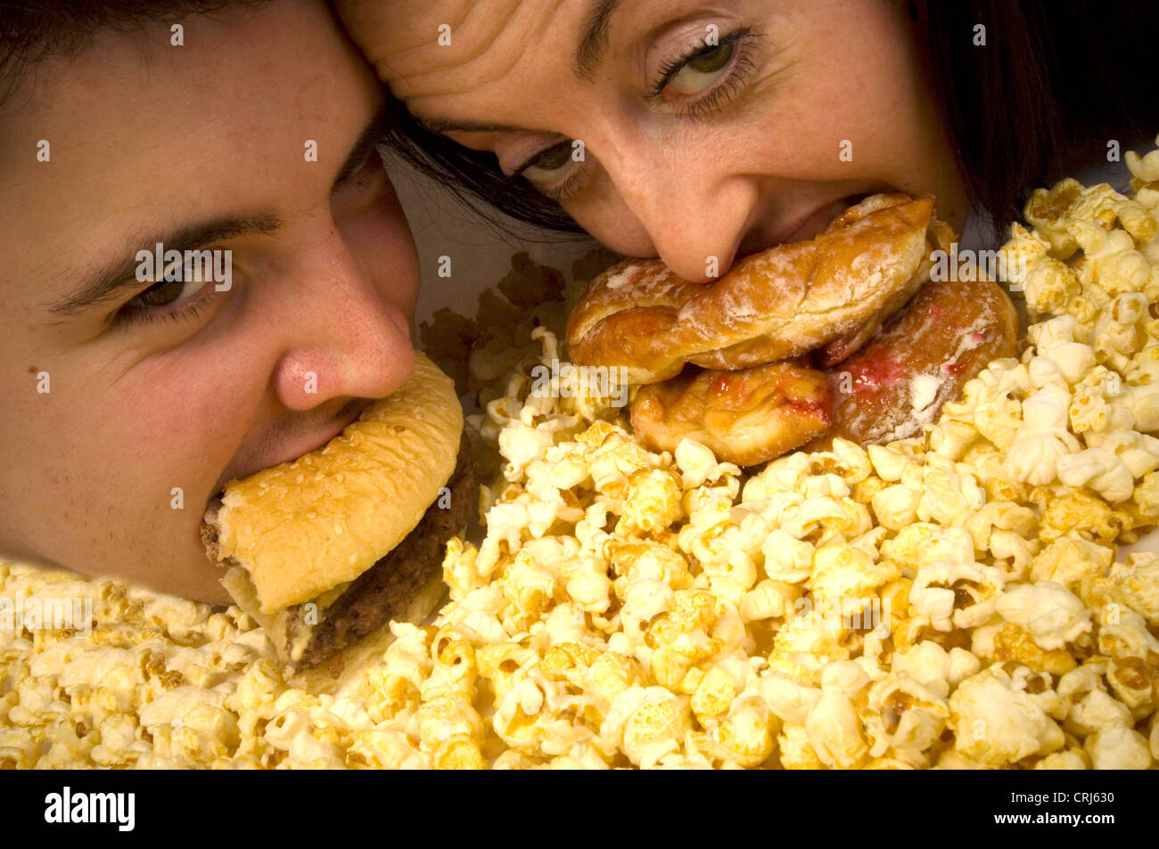 Ein junges Paar von Junk-Food abhängigen einen Hamburger und eine Marmelade-Donut mit einem Vorrat an Popcorn zu essen, zu folgen. Diese Lebensmittel, unter anderem sind von Junk-Food-Aktivisten im globalen Kampf gegen Fettleibigkeit als fehlerhaft gekennzeichnet mit schlechten Nährwert kritisiert worden. Der Begriff Â €˜junk Foodâ€™ wurde geprägt von Michael Jacobson, Direktor des Center for Science in öffentlichen Inte Stockfoto