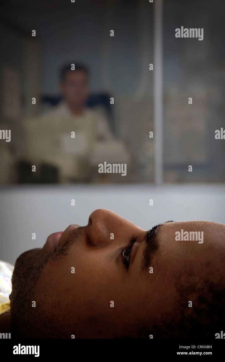 Ein Radiologe sitzen hinter Glas als Schutz gegen Radioaktivität, wie er ein Patient auf einem Bett liegend x-Strahlen. Stockfoto