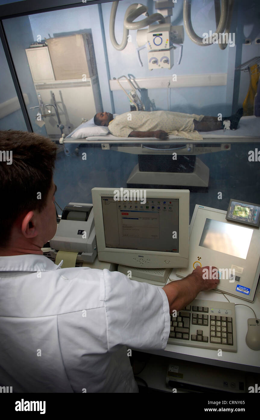 Ein Radiologe sitzen hinter Glas als Schutz gegen Radioaktivität, wie er ein Patient auf einem Bett liegend x-Strahlen. Stockfoto