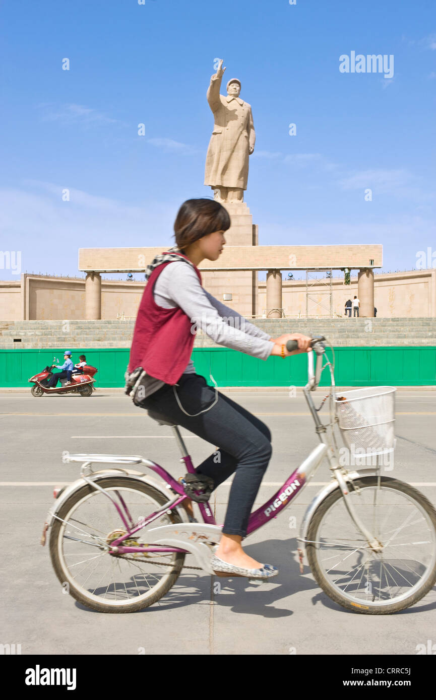 Mit Bewegungsunschärfe.  Eine junge Chinesin auf auf einem Fahrrad fahren Sie vorbei an der Statue von Mao Zedong in Kashgar. Stockfoto