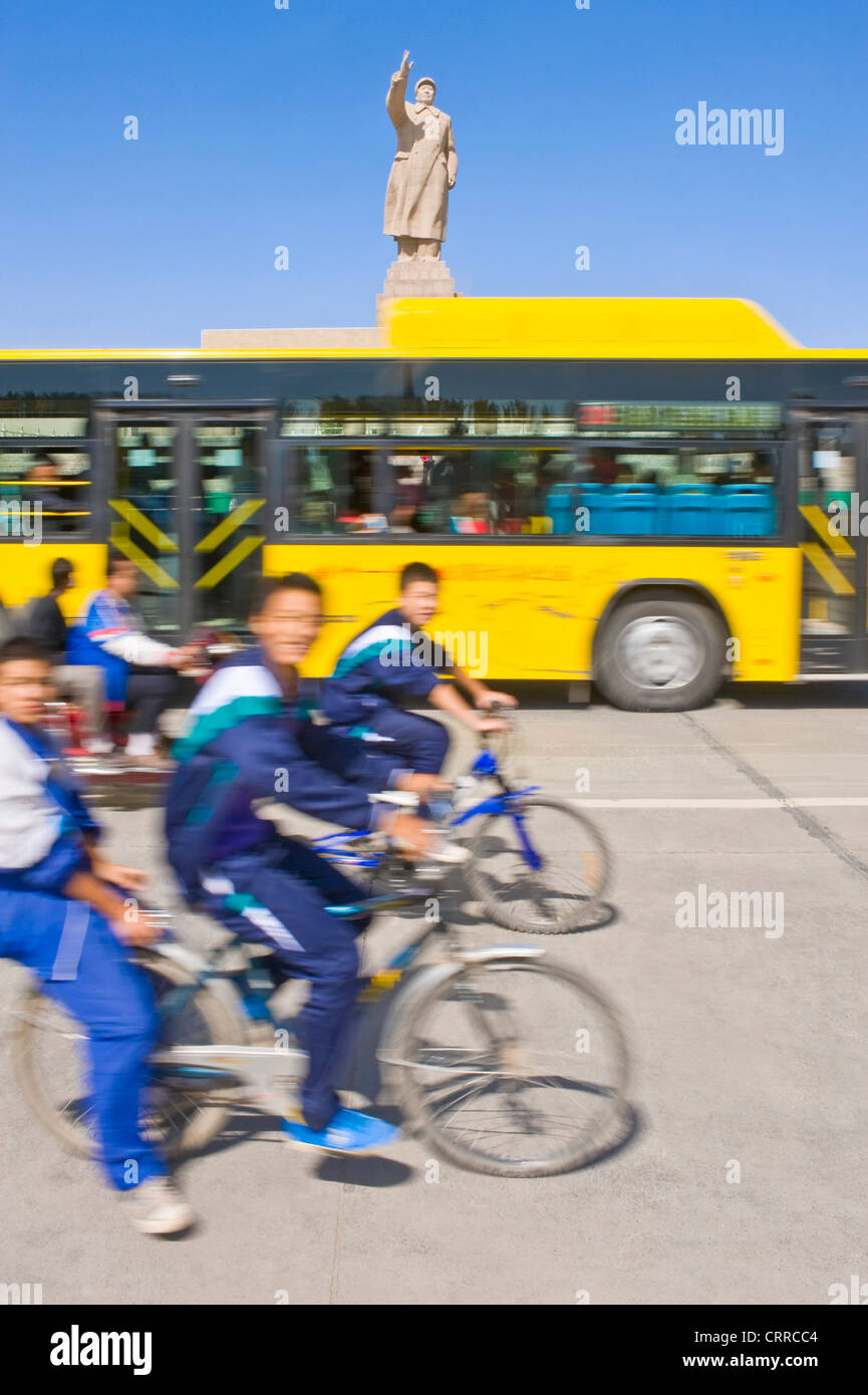 Mit Bewegungsunschärfe.  Junge chinesische Schuljungen auf Fahrrädern fahren Sie vorbei an der Statue von Mao Zedong in Kashgar. Stockfoto