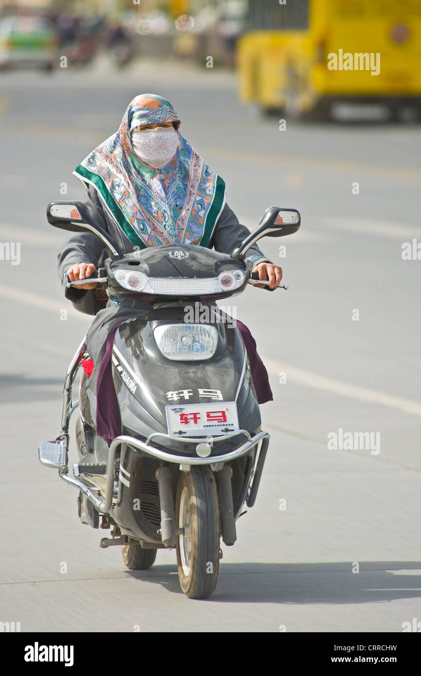Eine chinesischen uigurischen Frau reist entlang einer Straße auf ihrem Roller in Kashgar. Stockfoto