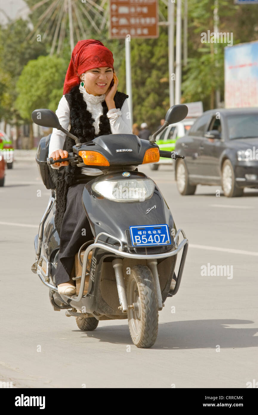 Eine chinesischen uigurischen Frau reist entlang einer Straße auf ihrem Roller beim Telefonieren mit ihrem Handy in Kashgar. Stockfoto