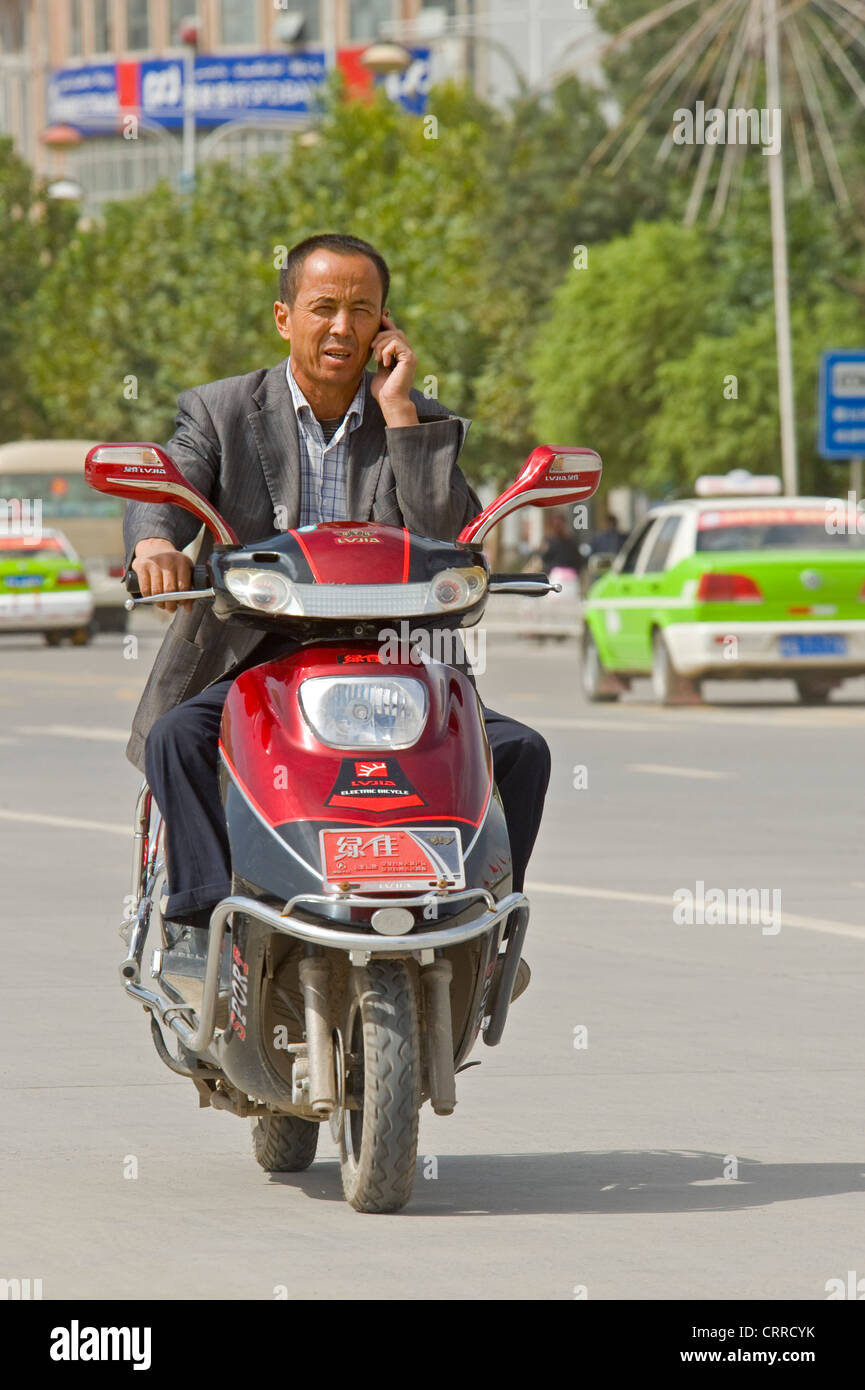 Ein chinesischer Mann reist entlang einer Straße auf seinem Roller während des Gesprächs auf seinem Handy in Kashgar. Stockfoto