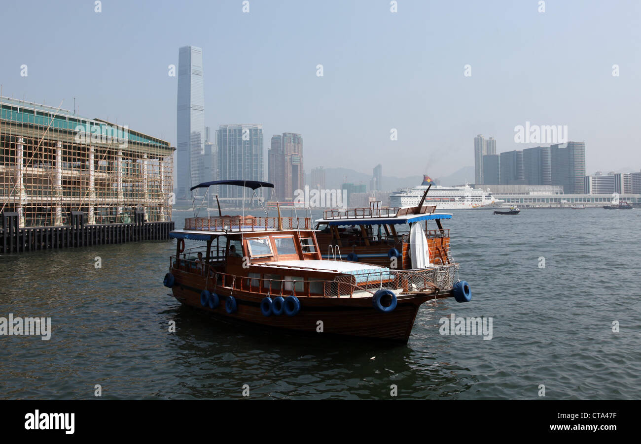 Es ist ein Foto von einem Boot in Hong Kong im Victoria Harbour. Wir sehen die Stadt im Hintergrund. Wir sehen auch eine Holz Dschunke Stockfoto