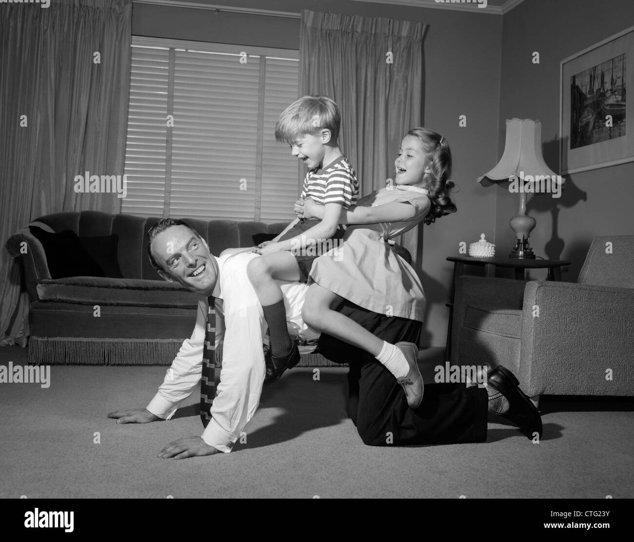 1950S 1960S HUCKEPACK PAPA TRAGEN JUNGE & MÄDCHEN AM BODEN DES WOHNZIMMERS Stockfoto