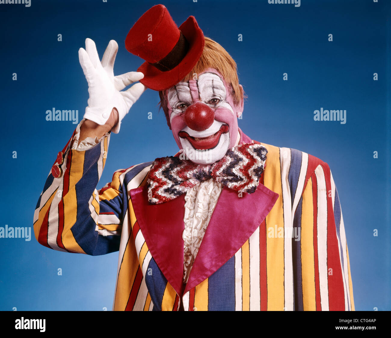 1970ER JAHRE PORTRAIT VON CLOWN IN GESTREIFTE KOSTÜM KLEINE ROTE ZYLINDERHUT KIPPEN Stockfoto