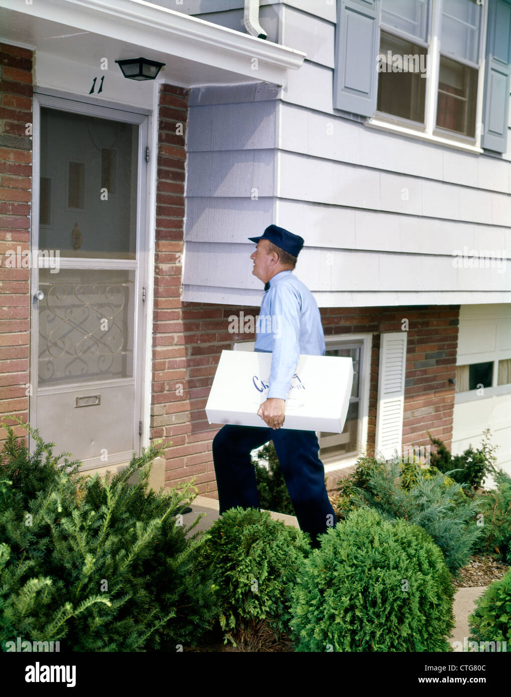 1960ER JAHRE DELILVERY MANN EINHEITLICHE HAUSTÜR VORSTADTHAUS LIEFERN GROßES PAKET SERVICE ARBEITNEHMER RETRO Stockfoto