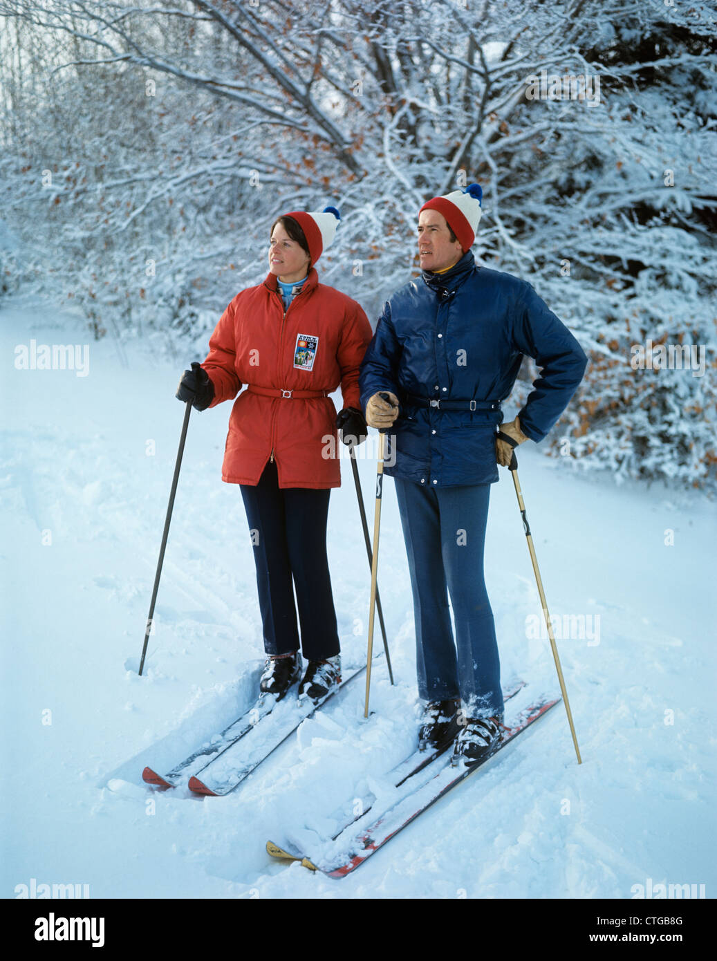 1970ER JAHREN VOLLER LÄNGE PAAR STEHEND AUF SKIERN FRAU ROTE JACKE MANN BLAUE KLEIDUNG RETRO Stockfoto