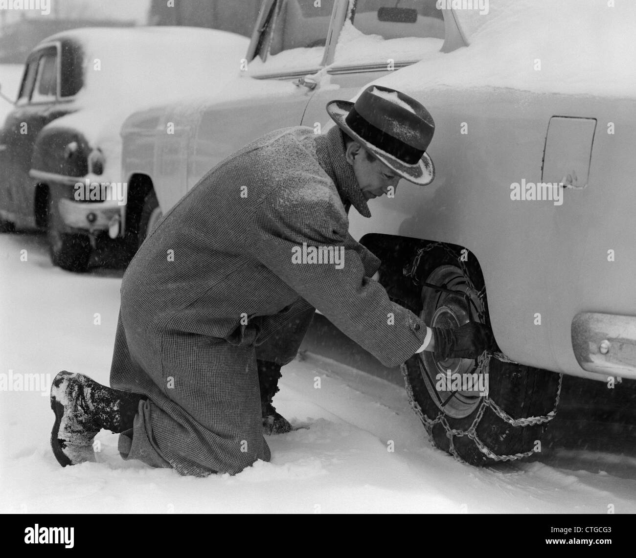 1950ER JAHRE MANN IN MANTEL & HUT KNIETE IM SCHNEE ANPASSEN AUF REIFEN-KETTEN Stockfoto