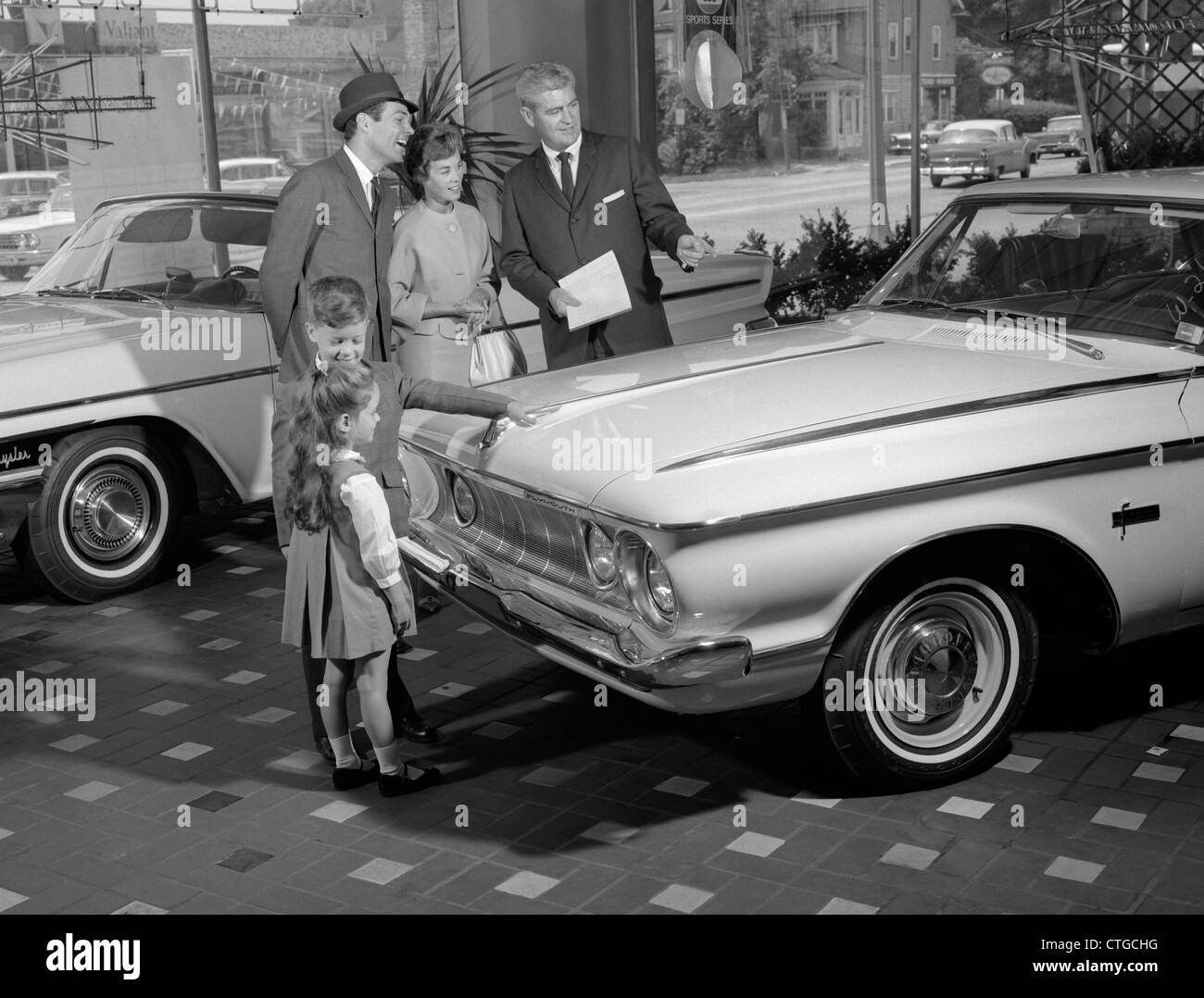 1960ER JAHRE FAMILIE 4 IM AUTO-SHOWROOM MIT VERKÄUFER SUCHEN, WEIßEN PLYMOUTH Stockfoto