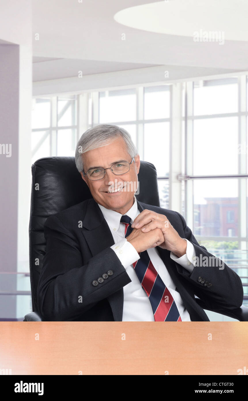 Lächelnd Mitte Alter Geschäftsmann mit seinen Händen zusammen sitzen an einem Schreibtisch in einem modernen Bürogebäude. Hochformat. Stockfoto