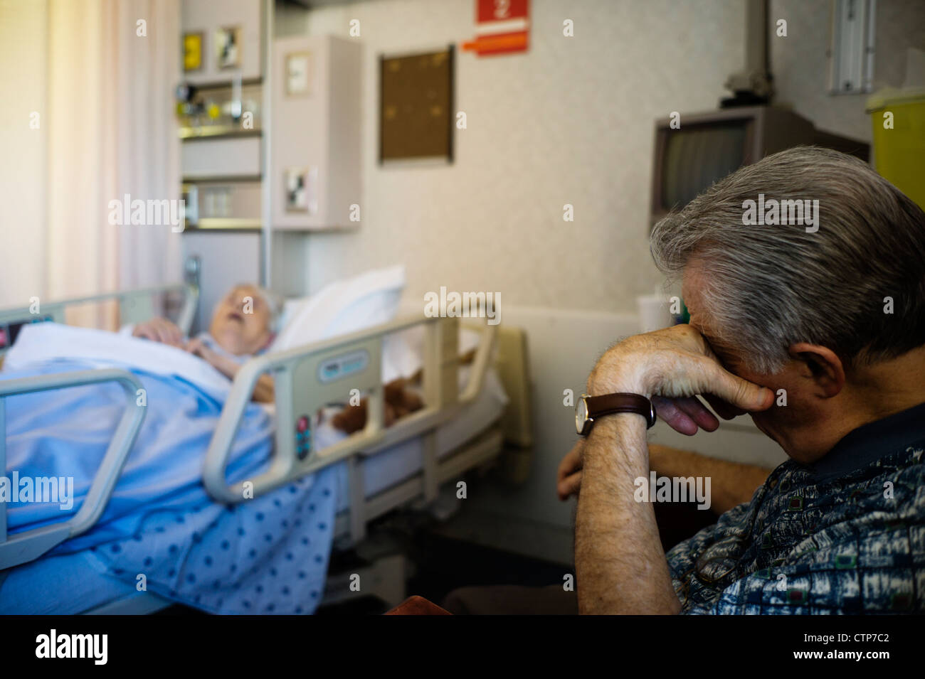 Alter Mann schlafend im Sessel mit alte Frau bewusstlos im Krankenhausbett im Hintergrund. Stockfoto