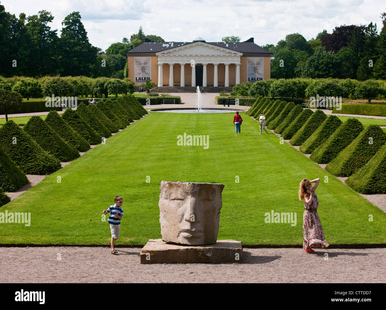 Eine Frau posiert für ein Foto, wie ein kleiner Junge um eine Statue im Botanischen Garten spielt. Uppsala, Schweden. Stockfoto