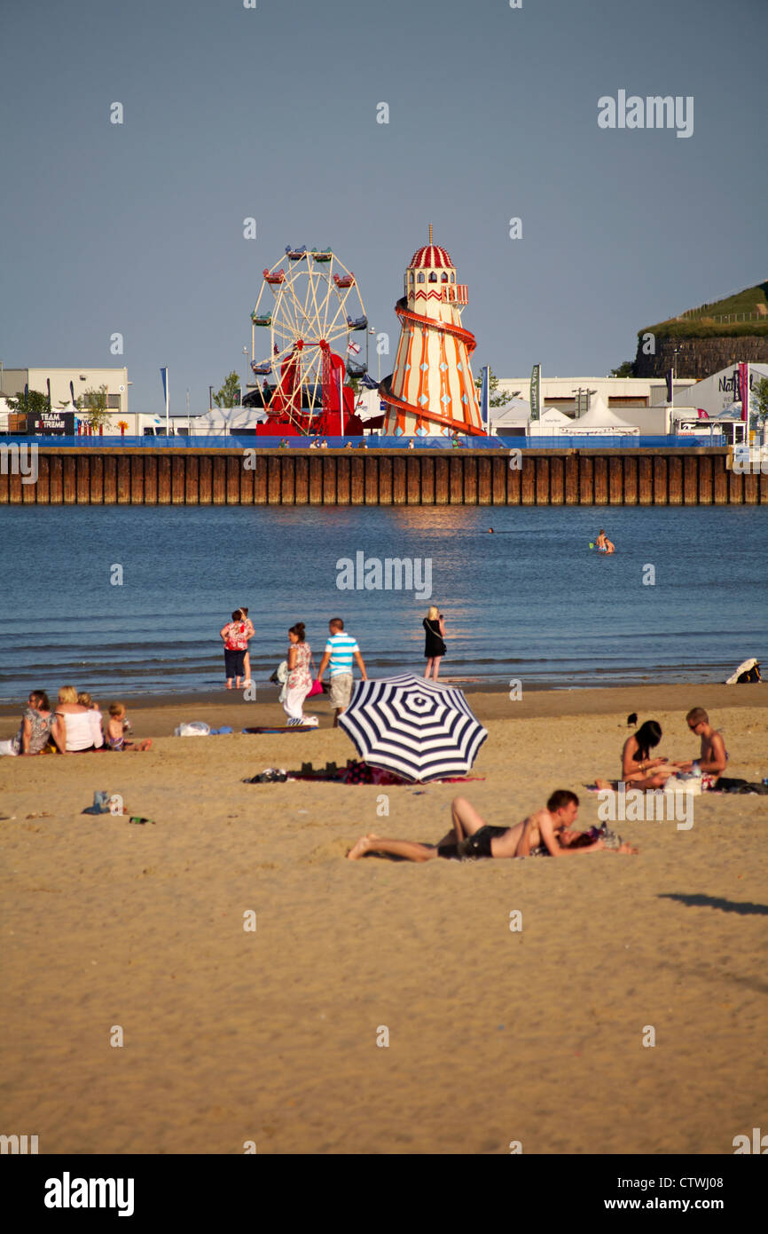 Nutzen Sie das warme Wetter am Weymouth Beach, Dorset UK im Juli mit den Fahrgeschäften auf dem Messegelände des Weymouth Bayside Festivals in der Ferne Stockfoto