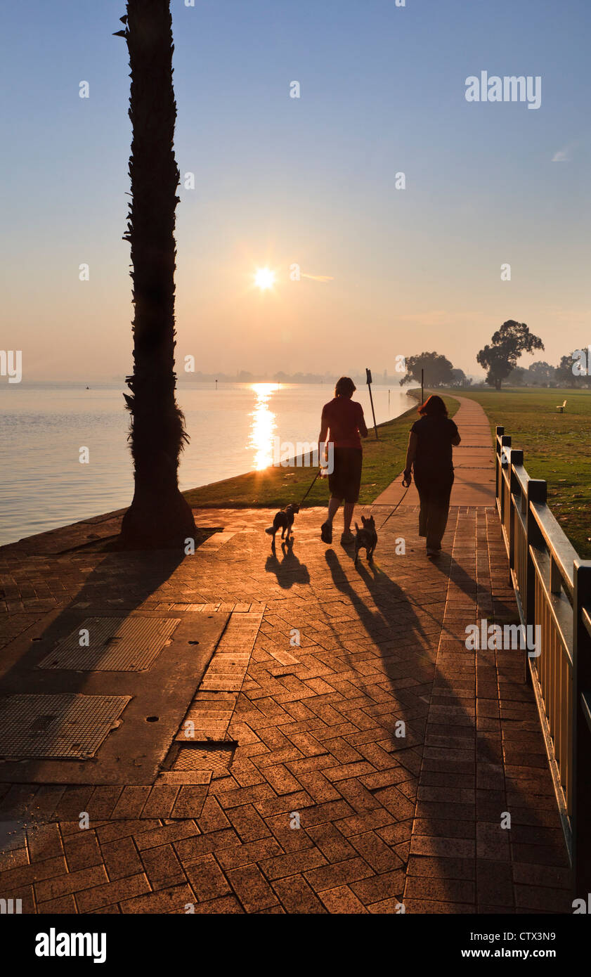 Zwei Frauen gehen Hunde bei Sonnenaufgang entlang einem Uferweg Stockfoto