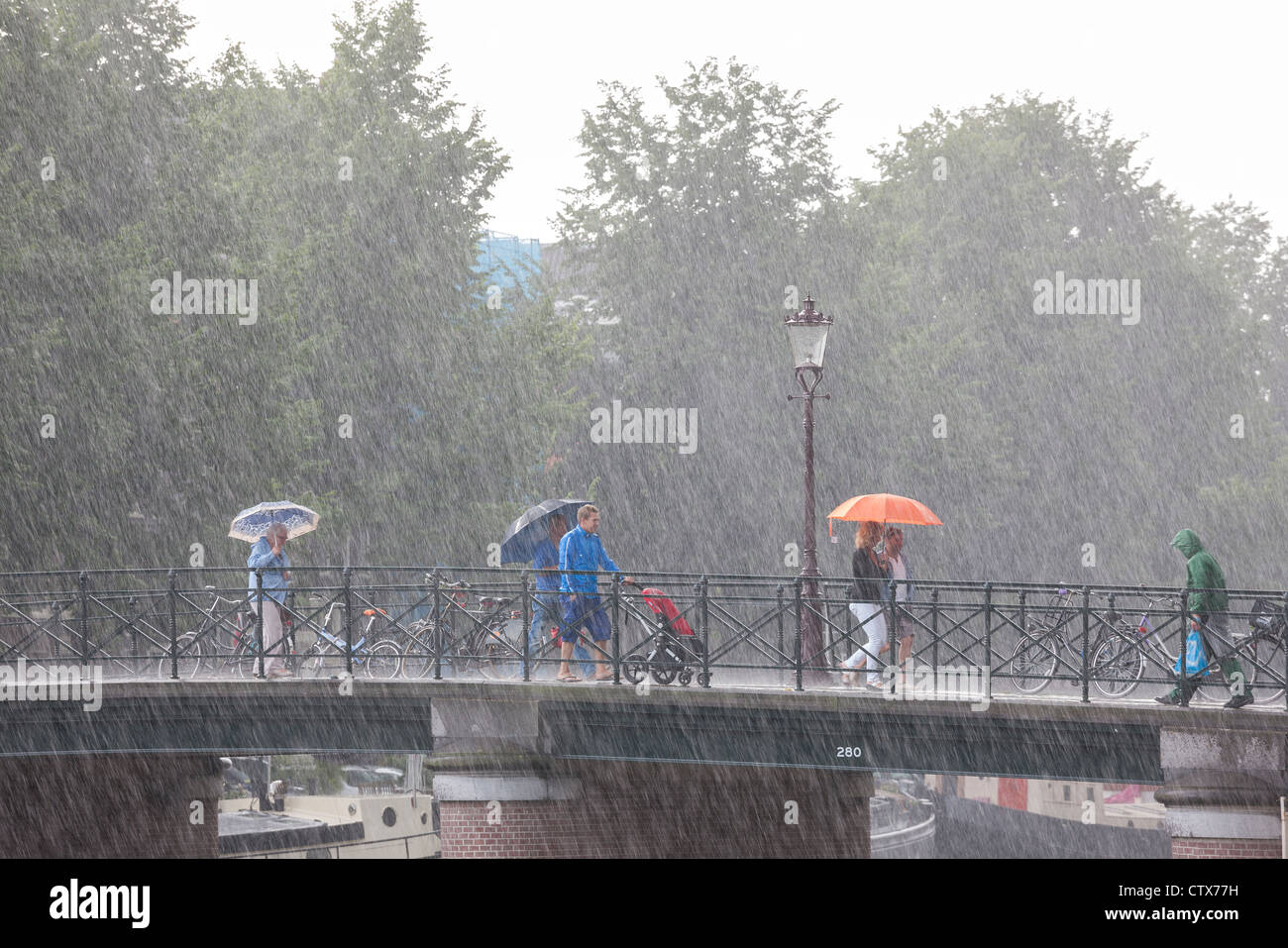 Amsterdam-Brücke. Plötzlichen reißenden Sommerregen. Menschen auf einer Brücke mit Sonnenschirmen. Eine lokale in Regenkleidung mit Einkaufstasche. Stockfoto
