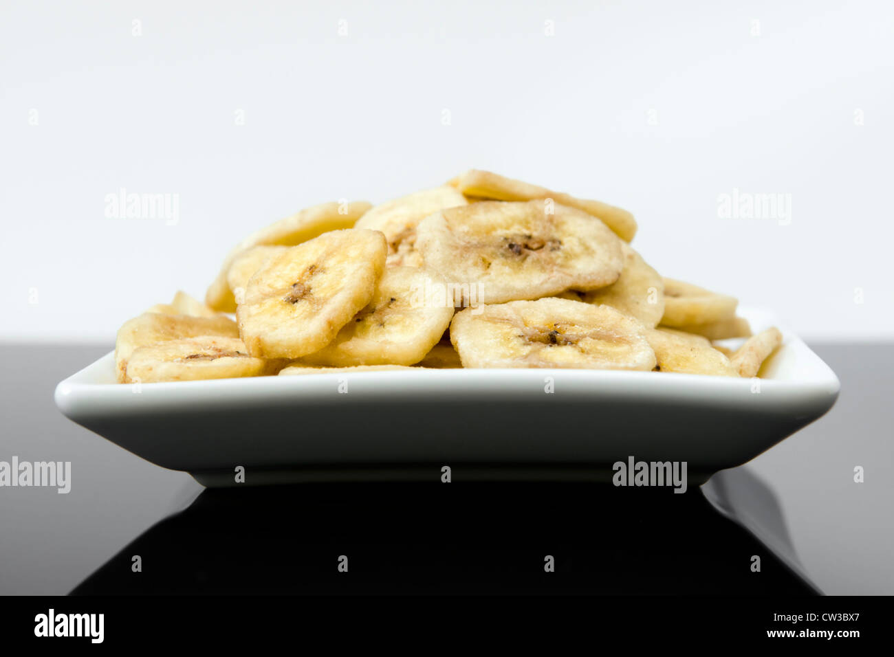 Getrocknete Bananenchips in weißer Schale in schwarz reflektierende Oberfläche vor einem weißen Hintergrund Stockfoto