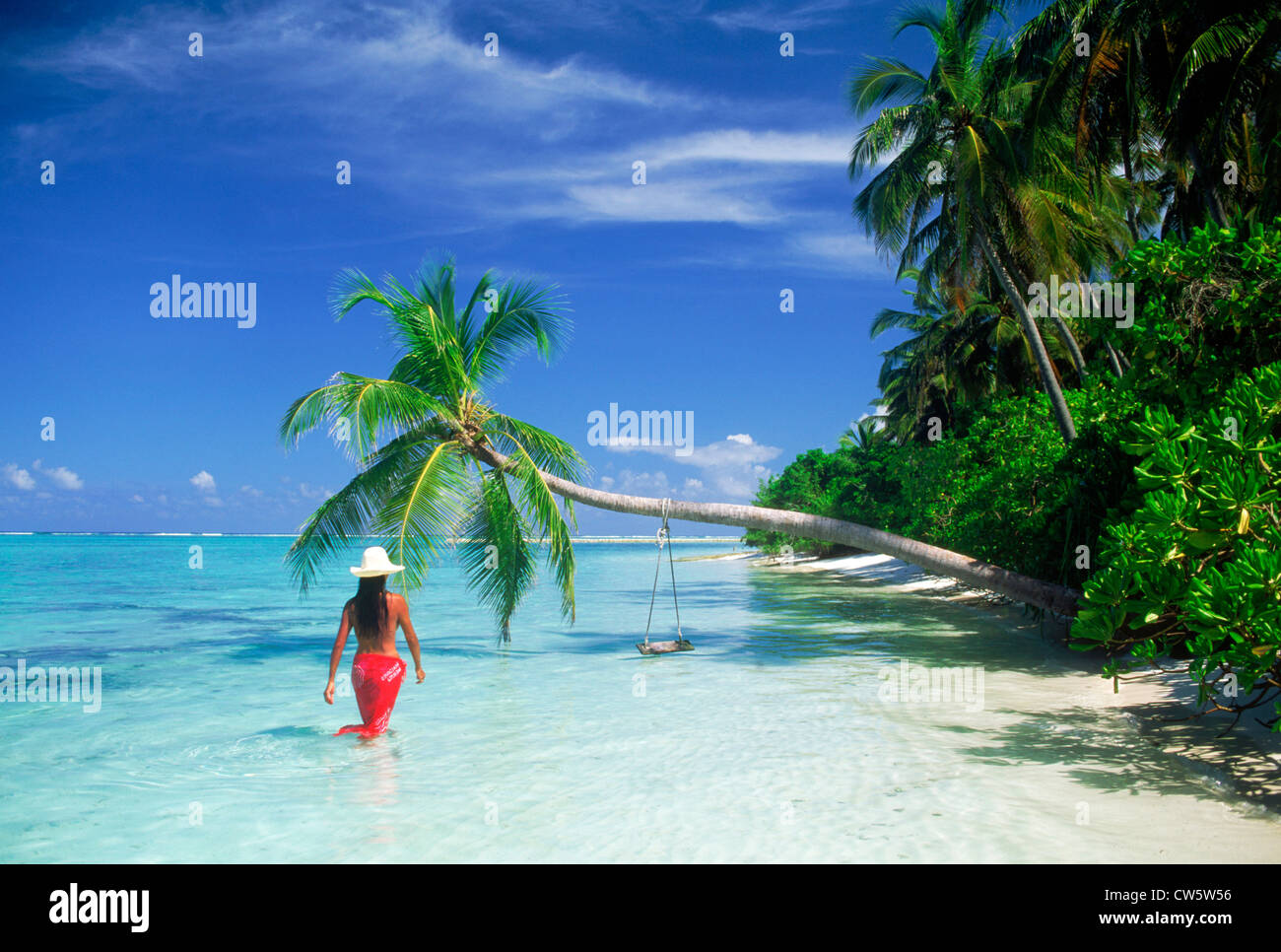 Frau trägt rote Sarong und weißen Hut Strand entlang mit überhängenden Palme inmitten der idyllischen tropischen Insel Urlaub Stockfoto