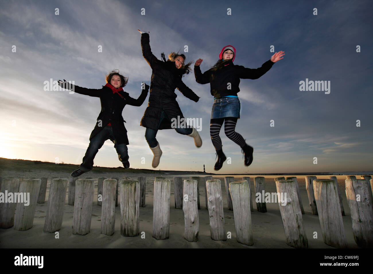 drei junge Frauen, die einen Sprung von Holzpfählen der Buhne am Strand, Niederlande, Zeeland, Breskens, Sluis Stockfoto