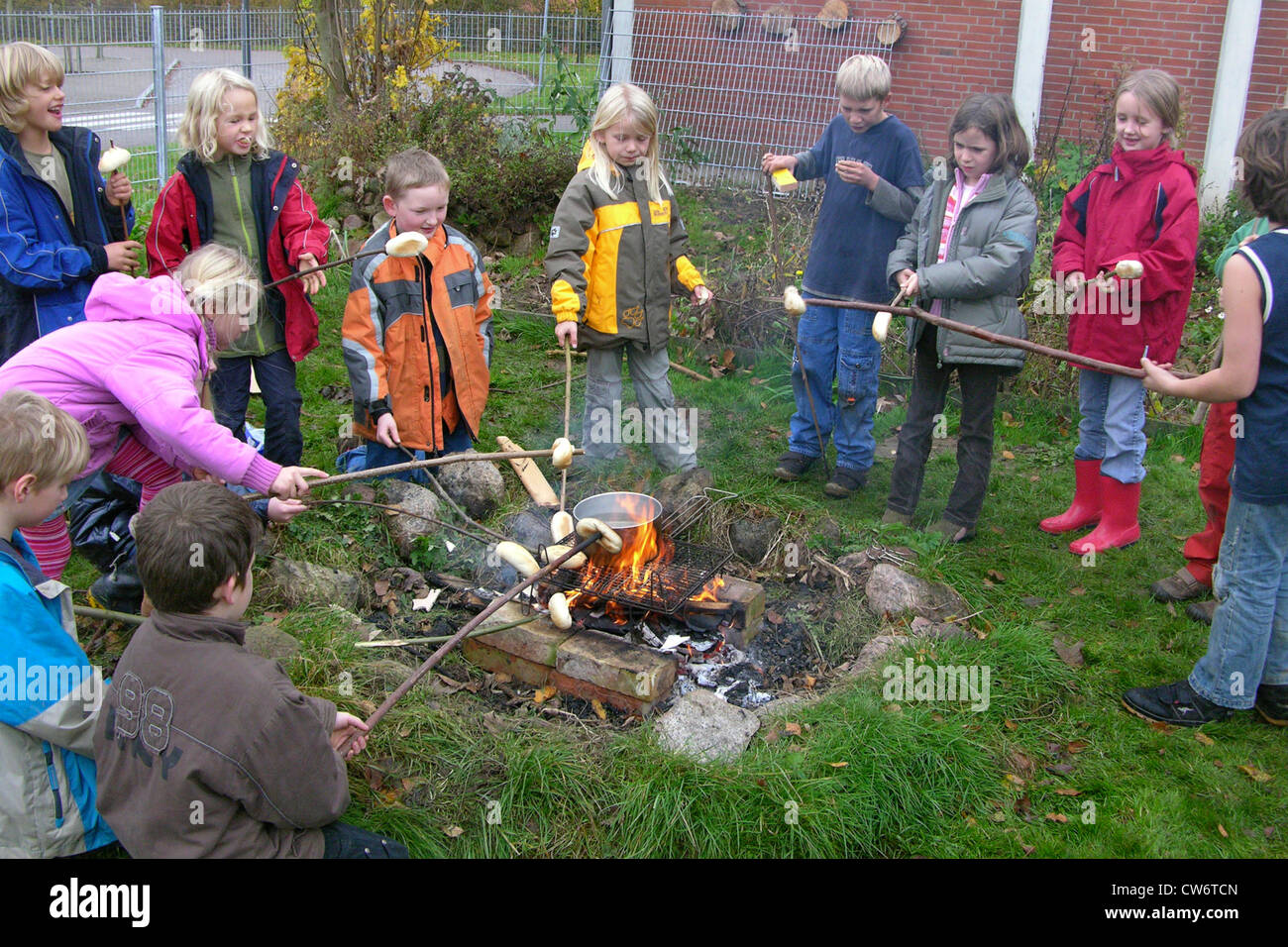 Kinder im Grundschulalter am Lagerfeuer Rösten Brot rollt auf Stöcke und kochende Wasser für Tee, Deutschland Stockfoto