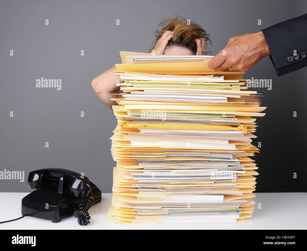 Eine ausgefranste weibliche Büroangestellte packt ihren Kopf in der Frustration, wie ihr Chef Haufen mehr auf ihrem Schreibtisch zu arbeiten. Stockfoto