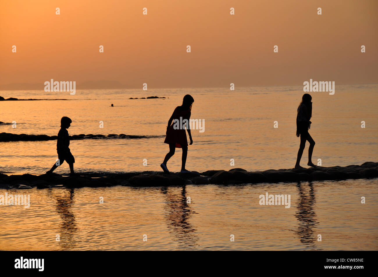 drei Kinder, die zu Fuß in Silouette am Strand bei Sonnenuntergang von links nach rechts Stockfoto