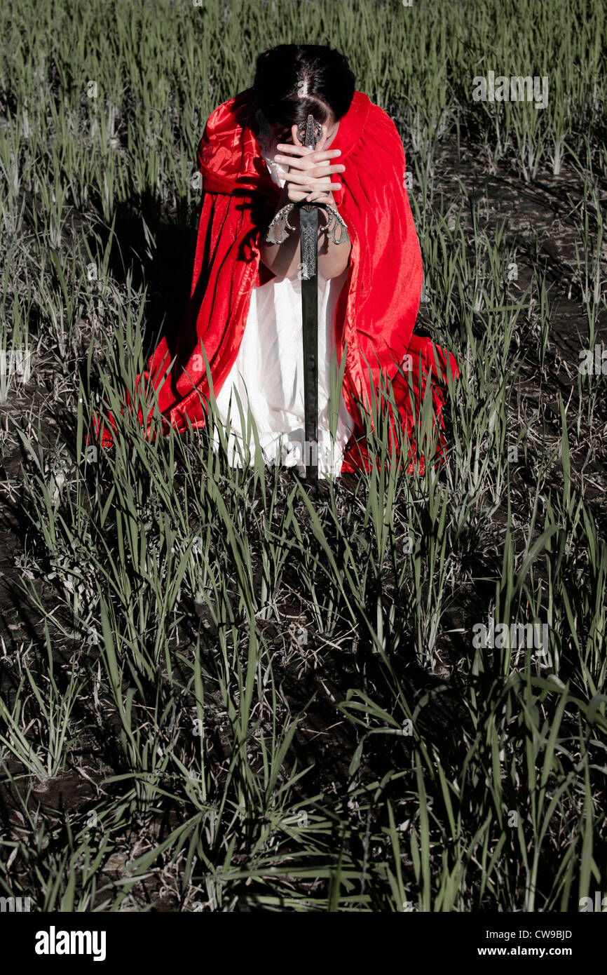 eine Frau in einem roten Mantel sitzt auf einem Feld mit einem Schwert Stockfoto
