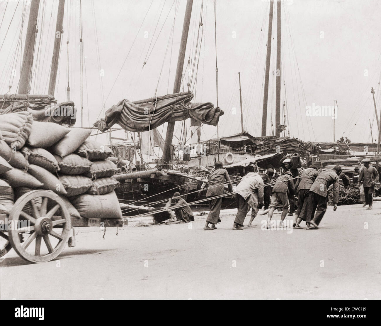 Bande von fünf chinesischen Dock Arbeiter Lean hart an Seilen, eine schwere Wagenladung von Säcken am Hong Kong Wasser zu schleppen. Zwischen Stockfoto