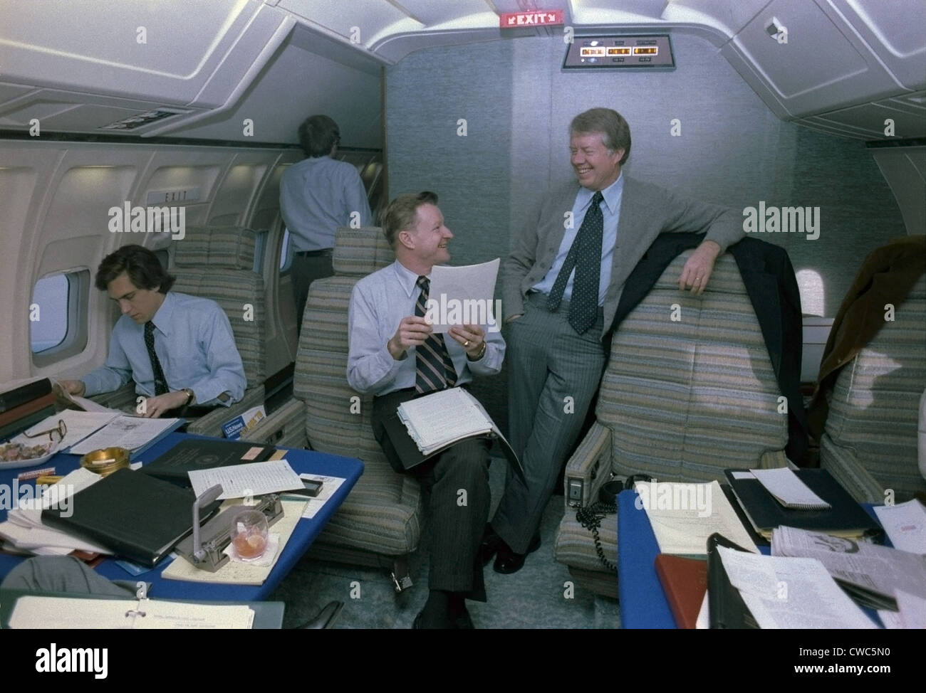 Zbigniew Brzezinski und Jimmy Carter in einem entspannten Gespräch an Bord der Air Force One, während eine junge Mitarbeiterin inmitten Papiere in arbeitet Stockfoto