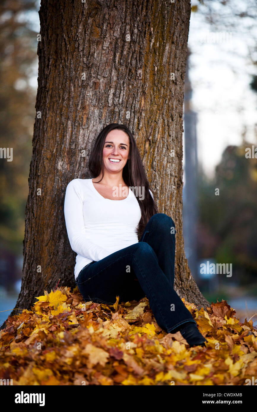 Eine schöne junge Frau lächelnd sitzen unter einem großen Baum im Herbst in Idaho. Stockfoto