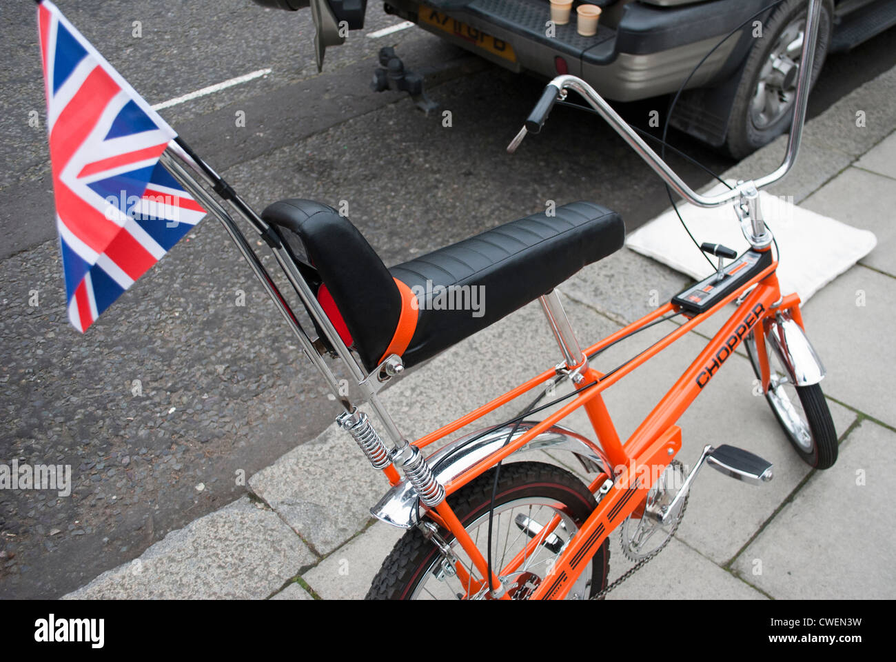 Orange-Chopper Fahrrad mit Anschluß-Markierungsfahne Wimpel geparkt auf Stein gekennzeichnet Bürgersteig neben Auto Stockfoto