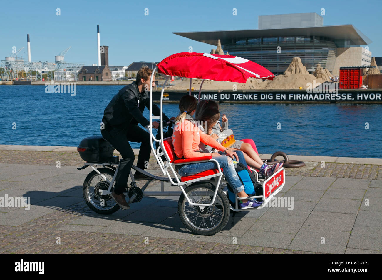 Fahrradkabine mit zwei Besichtigungstouristen in Larsens Plads, die das Royal Danish Opera House und Sandskulpturen am Ofelia Beach sehen. Kopenhagen, Dänemark. Stockfoto