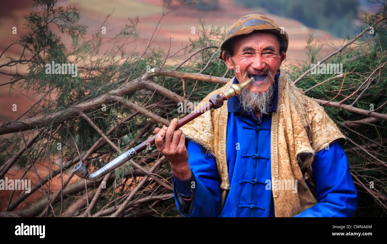 Ein lächelnder alter asiatischer Mann, der Pfeife raucht und auf seinem Rücken Äste trägt Stockfoto