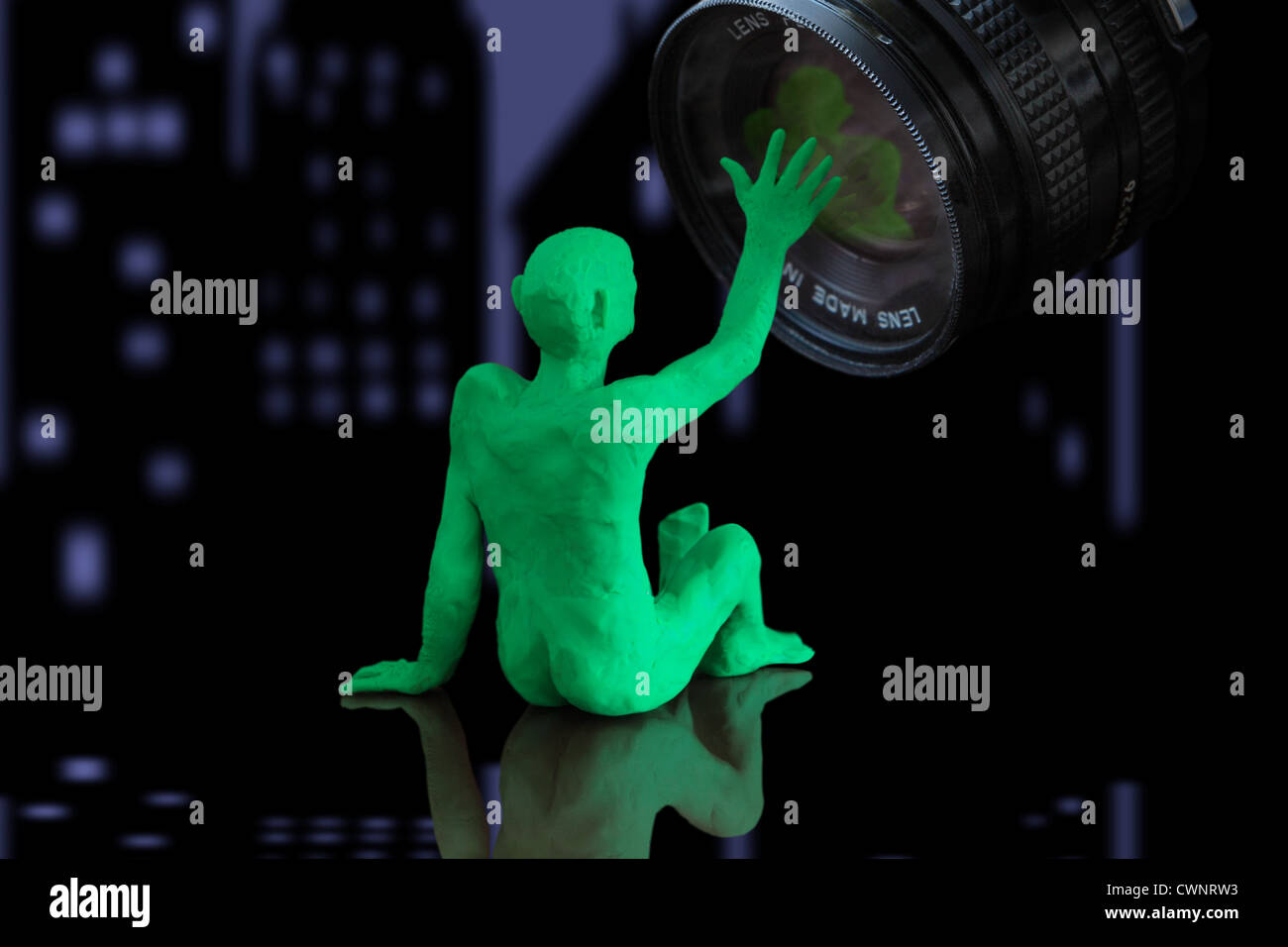 Wir sehen eine Figur, die eine Person darstellt, die sich hinsetzt und mit einer Kameralinse interagiert (Modelliermasse). Stadt bei Nacht im Hintergrund Stockfoto