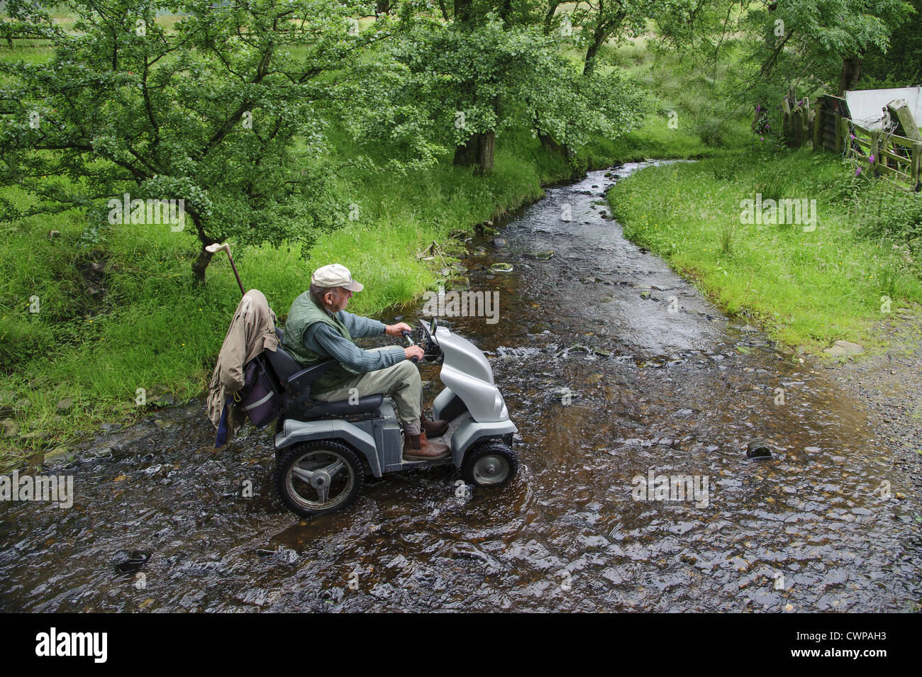 Älterer Mann mit Gehbehinderung mit "Tramper" (all Terrain Mobilität Scooter), um Bauernhof, zu besuchen über Bach bei Ford, Stockfoto