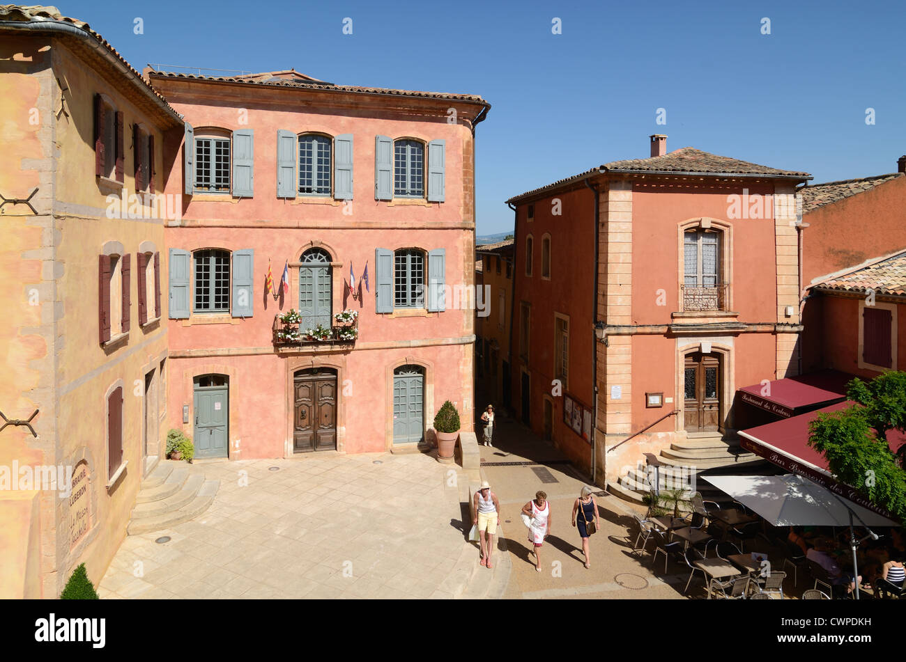 Touristen gehen über den Hauptplatz des Dorfes mit ocherfarbenen Gebäuden und dem Rathaus oder Mairie Roussillon Luberon Provence France Stockfoto