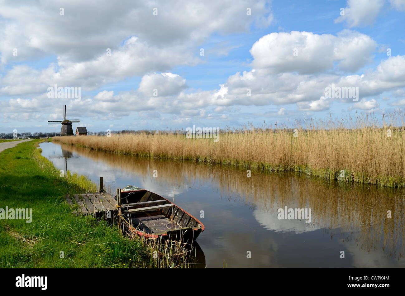 Typische Landschaft in Holland mit einem alten Boot in einem Graben mit einer Windmühle, Schilf und Wolken. Stockfoto