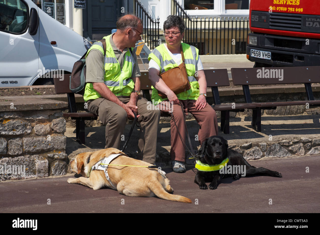 Blinden und sehbehinderten Mann und die Frau saß auf der Bank die Sonne zu genießen, mit Blindenhunden in Weymouth auf den Boden zu ihren Füßen gelegt Stockfoto