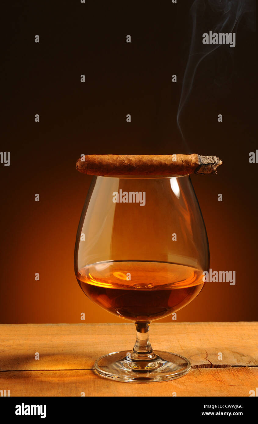 Schnaps Glas mit einer beleuchteten Zigarre auf seinen Rand über einen warmen Hintergrund. Rauch Weg weht von Zigarre. Stockfoto