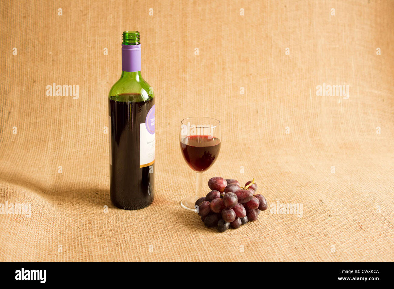 Flasche Rotwein mit einem Glas Wein, die Hälfte mit roten Trauben auf eine hessische Hintergrund gefüllt geöffnet. Stockfoto