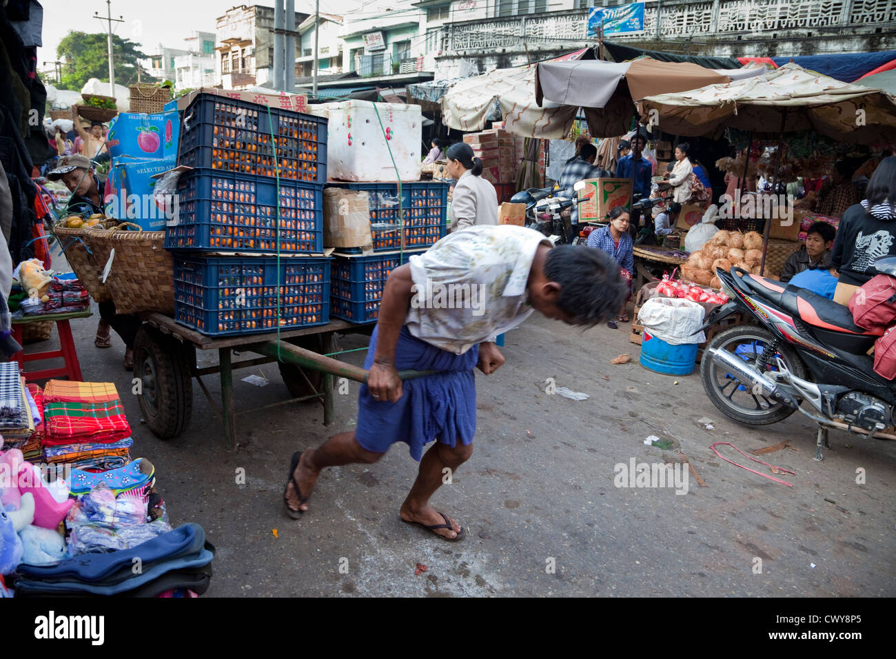 Myanmar, Burma. Mandalay-Markt-Szene. Mann kämpft um eine Last von Äpfeln und Orangen durch die Marktstraßen zu ziehen. Stockfoto